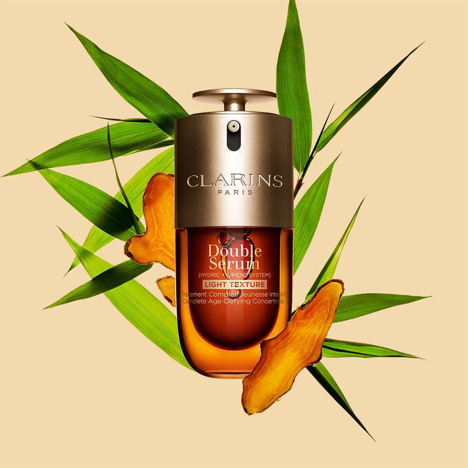 Un frasco de 30&nbsp;ml de Double Serum Textura Ligera de Clarins entre hojas de c&uacute;rcuma y ca&ntilde;a de Provenza frente a un fondo naranja.