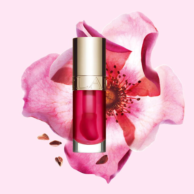 Foto del tono Pitaya de Lip Comfort Oil de Clarins junto a una flor de rosa mosqueta sobre un fondo rosa claro.