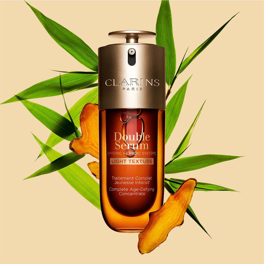 Un frasco de 70&nbsp;ml de Double Serum Textura Ligera de Clarins entre hojas de c&uacute;rcuma y ca&ntilde;a de Provenza frente a un fondo naranja.