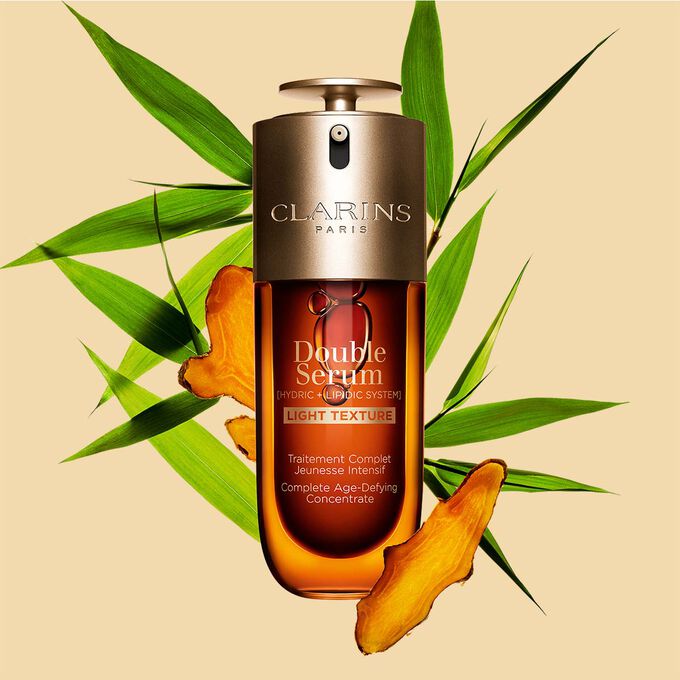 Un frasco de 70&nbsp;ml de Double Serum Textura Ligera de Clarins entre hojas de c&uacute;rcuma y ca&ntilde;a de Provenza frente a un fondo naranja.