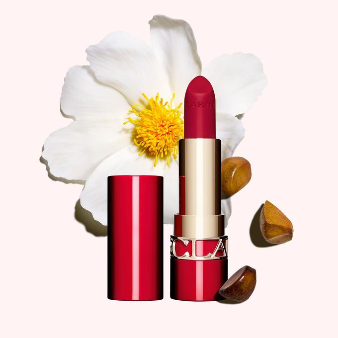 Foto del tono 742V de la barra de labios mate Joli Rouge de Clarins junto a una camelia y semillas de karité sobre un fondo rosa.