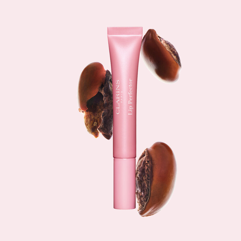 Tubo de Plumping Lip Perfector mostrado con semillas de karit&eacute; crudas para resaltar la intensa f&oacute;rmula de cuidado labial con efecto nutritivo profundo.