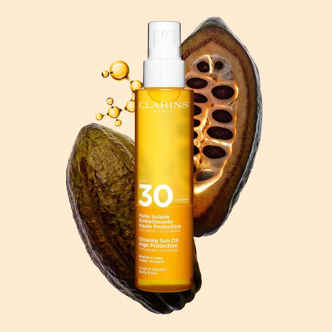 Foto del envase del spray naranja de aceite corporal y capilar SPF&nbsp;30 al lado de granos de cacao con un fondo naranja claro