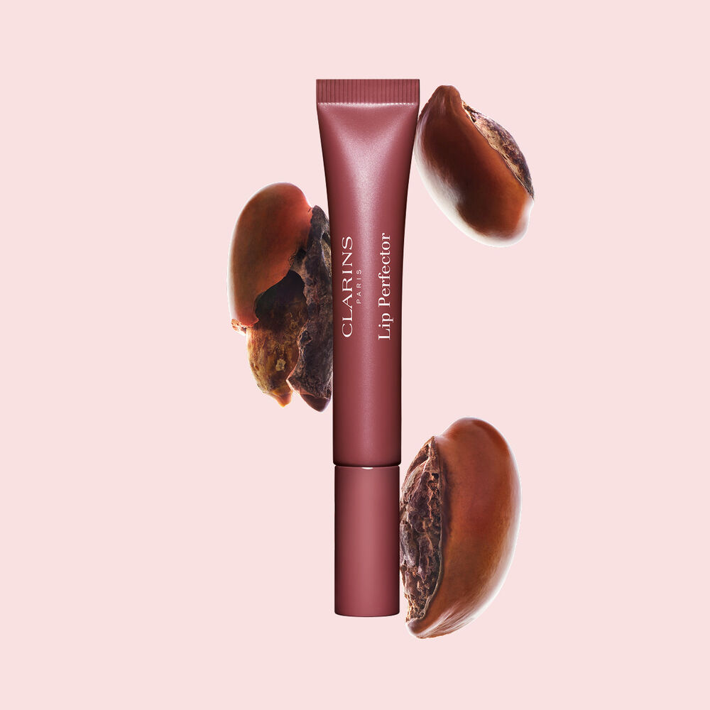 Tubo de Plumping Lip Perfector mostrado con semillas de karit&eacute; crudas para resaltar la intensa f&oacute;rmula de cuidado labial con efecto nutritivo profundo.